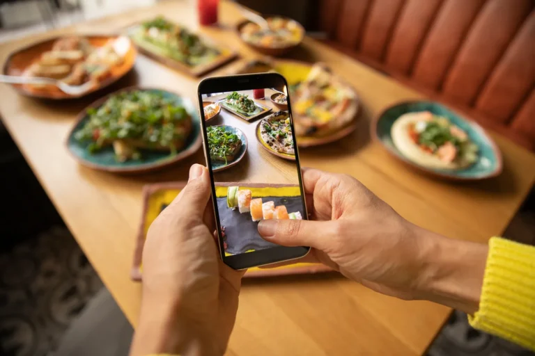 Mãos segurando o celular para fotografar pratos em uma mesa de restaurante