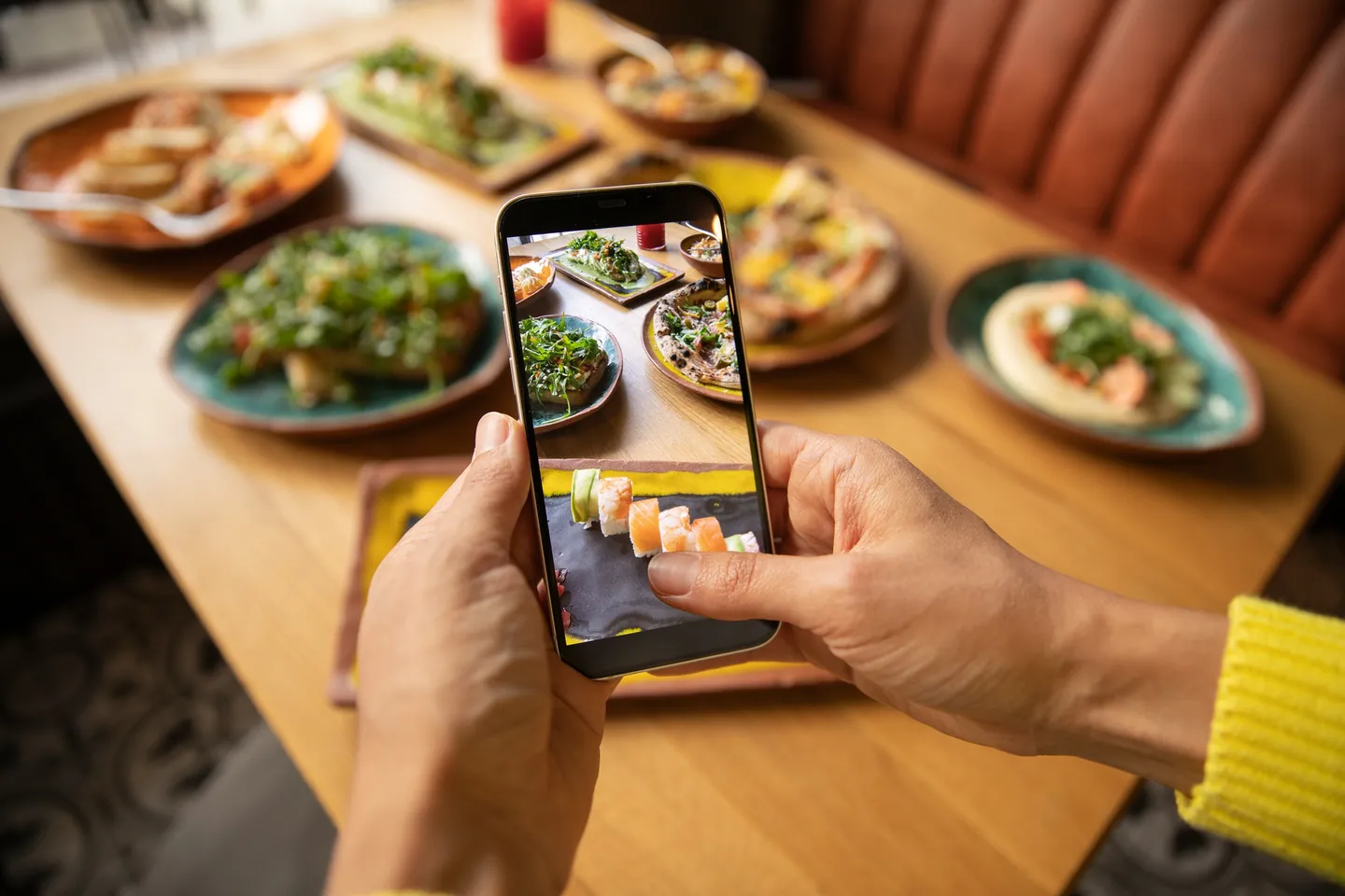 Mãos segurando o celular para fotografar pratos em uma mesa de restaurante